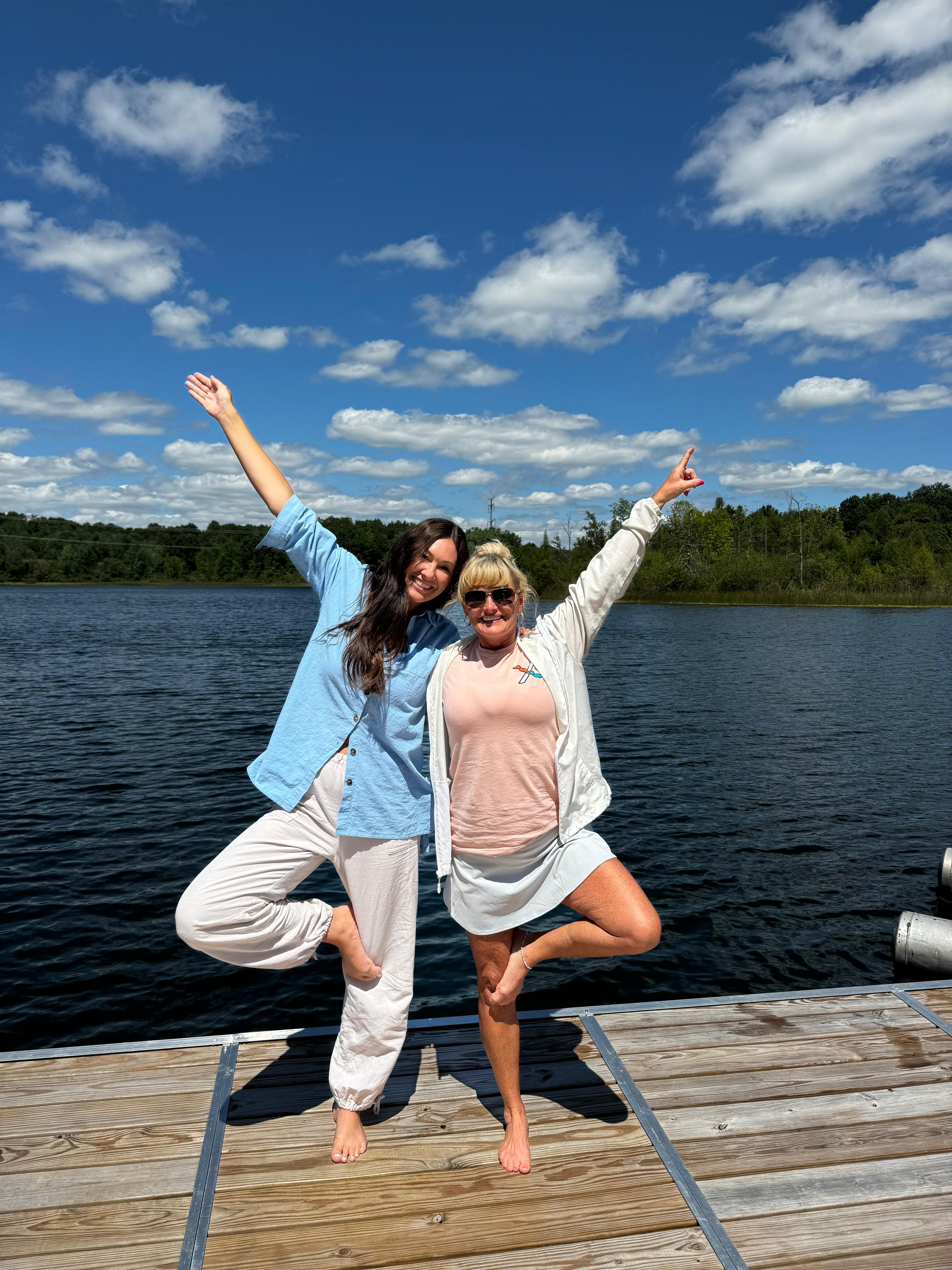 Two retreat guests in tree pose on the lake dock, arms raised under an open blue sky